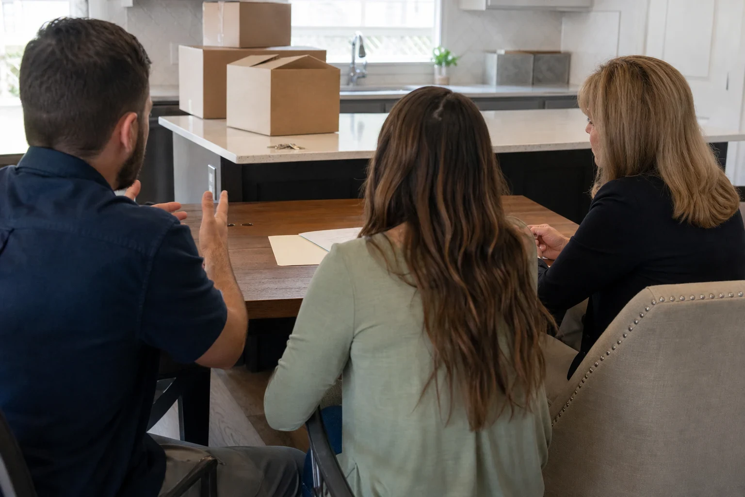 Agent with blonde hair listens as a young couple talks at a wooden kitchen table, with moving boxes and house keys on the counter behind them